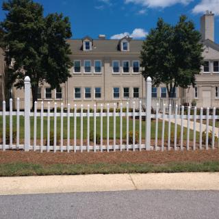 A view of a well-maintained building surrounded by a white picket fence and green grass under a clear blue sky.