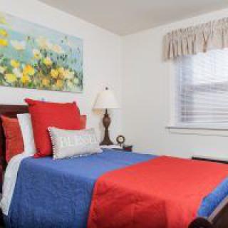 A cozy bedroom with colorful red and blue bedding, a decorative pillow, and a floral painting on the wall.