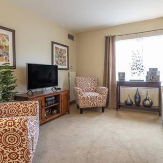 Cozy living room with patterned armchairs and a TV stand, softly lit by a large window.