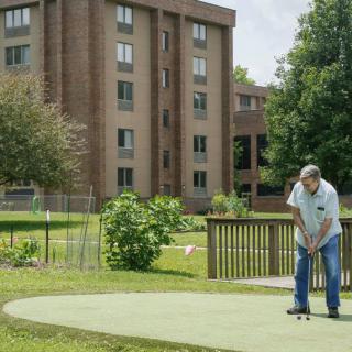 A man playing golf on a small putting green near a residential building.