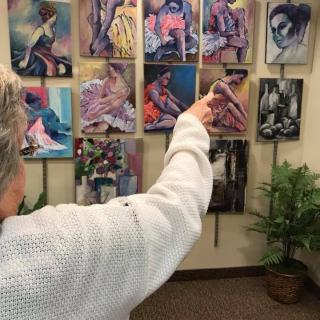 A person in a white sweater points towards paintings displayed on a wall in an art gallery.