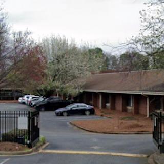 A view of a parking lot near a single-story building, surrounded by trees under a cloudy sky.