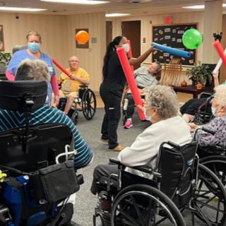 A group of seniors engage in a lively indoor activity with balloons and pool noodles in a nursing home setting.