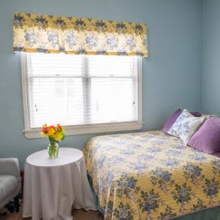 A cozy bedroom featuring light blue walls and floral bedding, illuminated by natural light from the window.