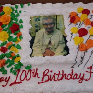 A beautifully decorated cake celebrating a 100th birthday with vibrant flowers, balloons, and a photo.