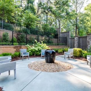 A serene outdoor patio area with chairs surrounding a central fire pit, set against a lush garden backdrop.