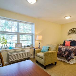 A cozy bedroom with a bed, armchair, and large window letting in natural light.