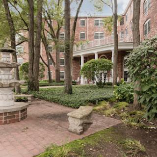 A serene garden area featuring a brick building backdrop and a charming fountain surrounded by lush greenery.