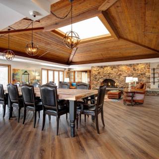 A spacious dining room with a wooden ceiling, skylight, and stone fireplace, featuring leather chairs and a hardwood floor.