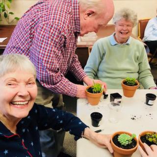 A group of seniors enjoying a gardening activity indoors, each holding a small potted plant and smiling together.