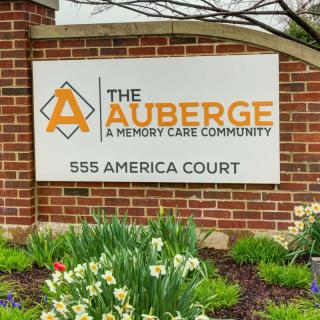 Sign for The Auberge, a memory care community, at 555 America Court surrounded by landscaped flowers.