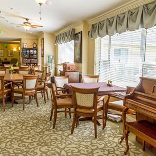 A cozy dining room with tables and chairs, featuring a piano and large windows with curtains.