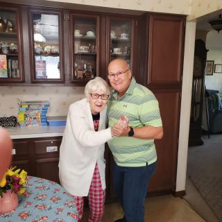 An elderly woman and a man posing together in a kitchen, both smiling.
