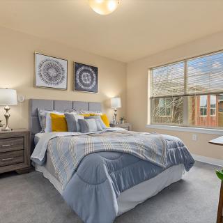 A cozy bedroom featuring a neatly made bed with blue and yellow accents, flanked by two bedside tables and lamps, with decorative artwork on the walls and a large window providing natural light.