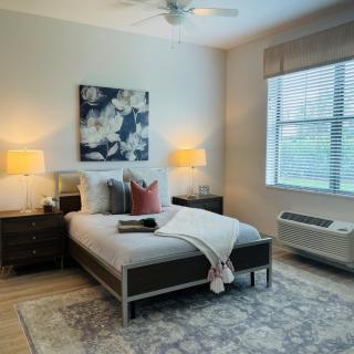 A cozy bedroom with a neatly made bed, bedside lamps, a large window, and decorative painting on the wall.