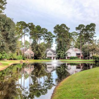 A scenic view of houses by a tranquil lake with a fountain and surrounding trees.