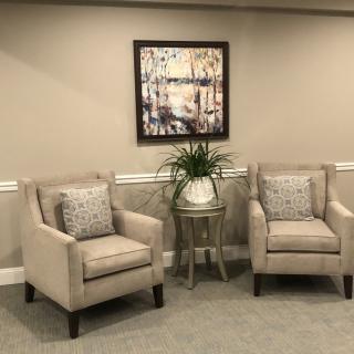 Two beige armchairs with cushions, a small side table with a plant, and framed artwork on the wall create a cozy living room setting.