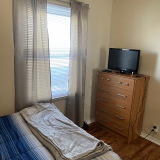 A cozy bedroom with a bed next to a window, featuring light curtains, wooden furniture, and a television on top of a chest of drawers.