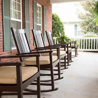 A row of wooden rocking chairs lined up on a spacious porch with a brick exterior.