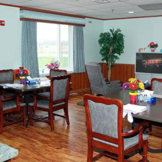 A well-furnished dining room with wooden floors and elegant chairs around tables, decorated with flowers and surrounded by large windows and a potted plant.