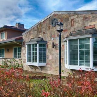A charming house with a stone facade and large windows, surrounded by landscaped plants under a cloudy sky.