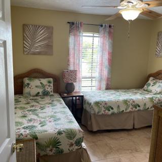 A cozy bedroom with two twin beds featuring floral bedding, complemented by a ceiling fan and soft natural light from the window.