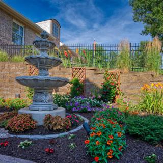 A beautifully landscaped garden featuring a multi-tiered fountain surrounded by vibrant flowers and a brick retaining wall.
