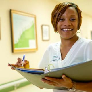 A smiling nurse holding a clipboard in a hospital corridor.