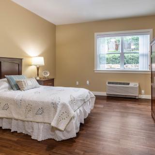 A cozy bedroom with a bed, nightstand, lamp, and a large window letting in natural light.