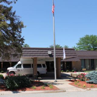 A view of a single-story building with an American flag, surrounded by well-maintained landscaping and a transportation van parked nearby.