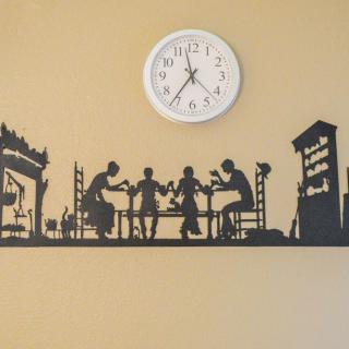 A wall featuring a round clock above a black silhouette sticker of people dining, set against an urban backdrop.