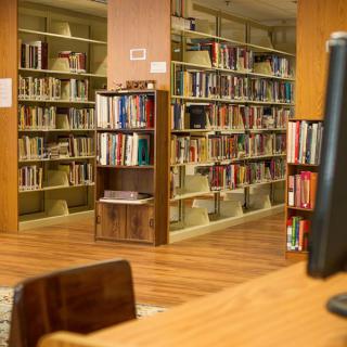 A quiet library setting with bookshelves filled with various books and a computer workstation in the foreground.
