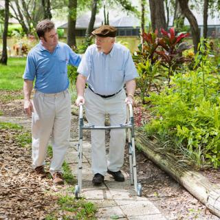 An elderly man using a walker is assisted by a caregiver as they walk together in a park.