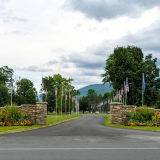 A scenic view of a road entrance framed by trees and flags, with mountains in the background under a cloudy sky.