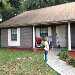 A group of people gathered on the porch of a house with a lawn and trees.