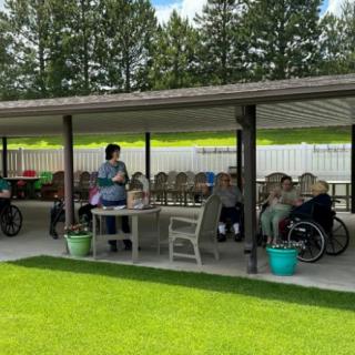 A group of people gathered under a covered patio with tables and chairs, surrounded by greenery.