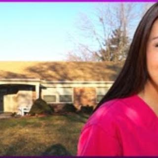 A woman in pink scrubs smiles warmly in an outdoor setting, illuminated by sunlight.