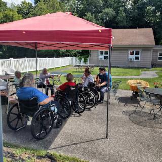 A group of senior citizens enjoying a sunny day outside, gathered under a red tent in their wheelchairs.
