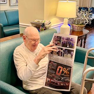 An elderly man sits comfortably in a chair, reading a newspaper under the warm glow of a lamp.