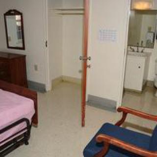A simple room with a bed, chair, mirror, open closet, and a sink area.