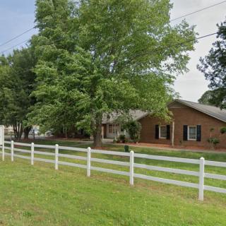 A brick house with a white fence and a large tree in front, surrounded by a green lawn.
