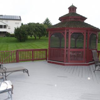 A wooden gazebo on a spacious deck with outdoor furniture, overlooking a green lawn.