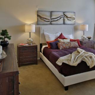 A stylishly decorated bedroom featuring a large bed with multiple pillows and a throw blanket, flanked by two nightstands with lamps and artwork above the headboard.