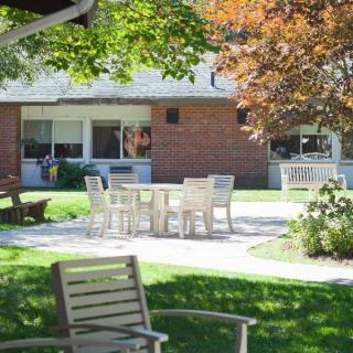 A peaceful garden patio area with chairs and tables, surrounded by greenery and a brick building.