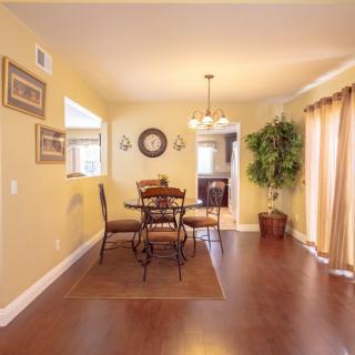 A cozy dining room with wooden flooring, furnished with a table and chairs beneath a chandelier. The space is decorated with wall hangings and features a large indoor plant by the window.