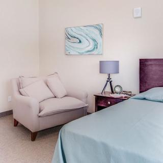A cozy bedroom corner featuring a plush chair, a bedside table with a lamp, and serene wall art.