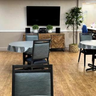 A cozy room with tables and chairs, featuring a television on the wall and decorative plants.