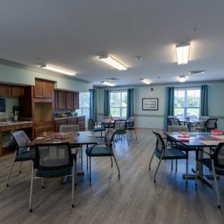 A community dining room with multiple tables and chairs, featuring a kitchen area and large windows.