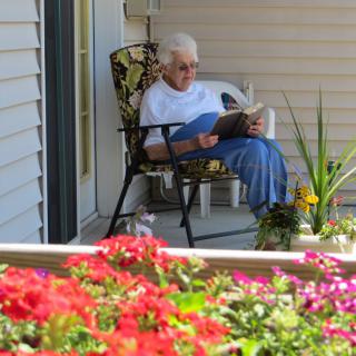 An elderly person sitting on a porch chair, reading a book surrounded by colorful flowers.