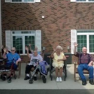 A group of elderly people waving while sitting outside in front of a building.